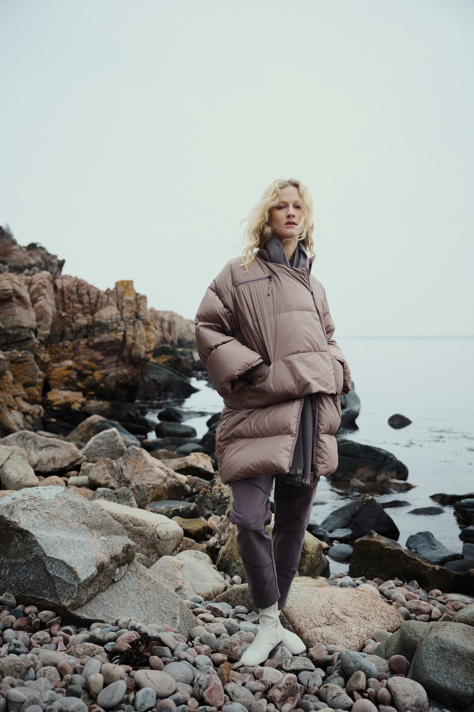 Model in een zachte, sculpturale jas aan een rotsachtige Scandinavische kust