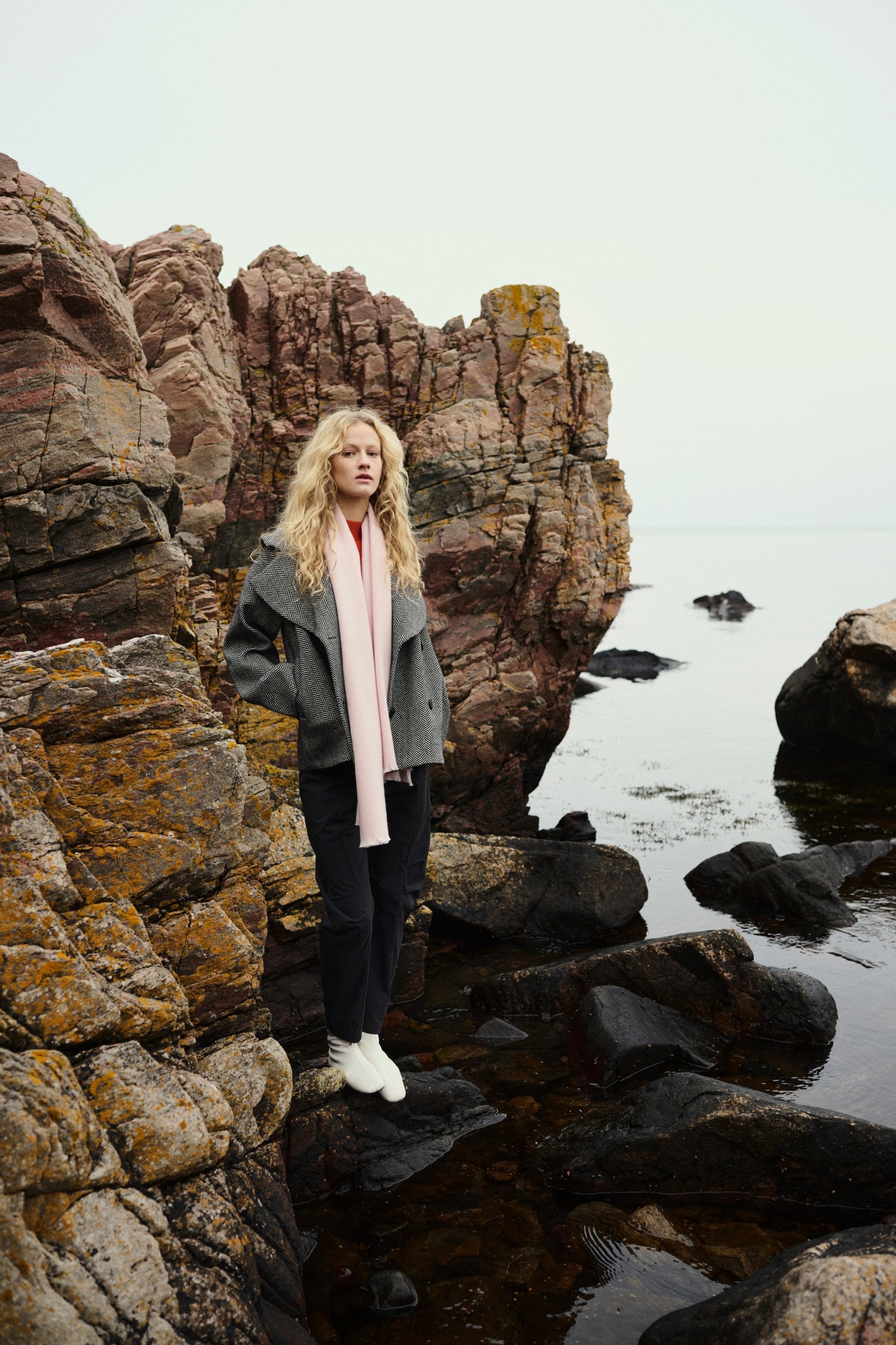 Model in een getailleerde visgraatjas en lichte sjaal tussen rotsen aan de kust